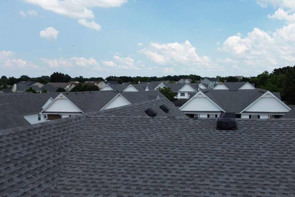 Aerial view across gray shingle roofs toward rows of matching white-gabled residential buildings under a partly cloudy sky.