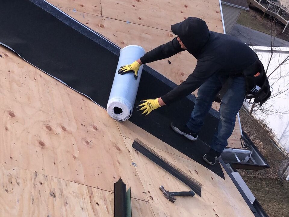 Roofer in a dark hooded sweatshirt and yellow work gloves rolling a length of black roofing underlayment across a plywood roof deck, with metal trim pieces and a hammer nearby.