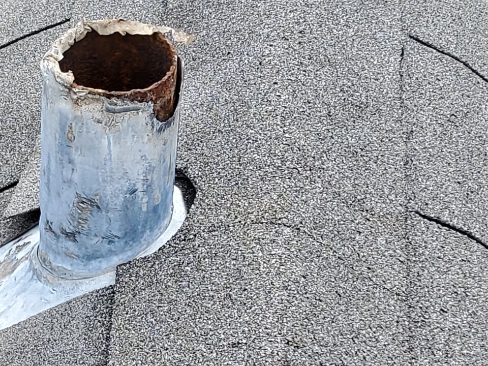 Vent pipe with a torn, jagged metal flashing sleeve at its top and rust inside the pipe, on a gray shingle roof.