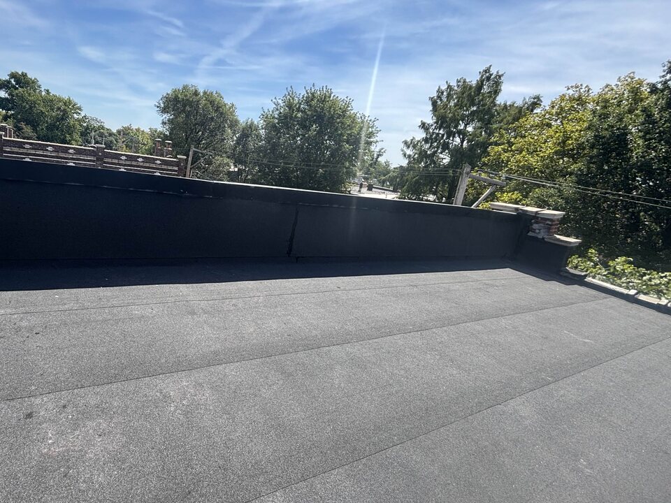 Flat roof covered in dark gray granulated modified-bitumen cap sheet, with black membrane running up a long parapet wall and trees against a blue sky beyond.