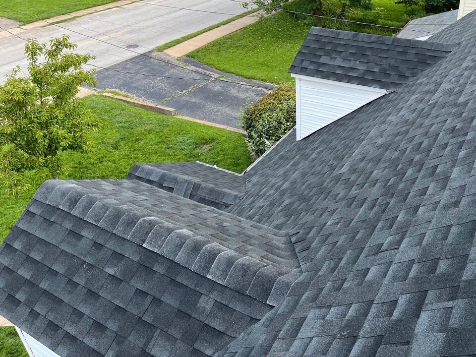 Dark gray shingle roof with a small dormer and capped roof lines, seen from above, with a lawn, a driveway and a street below.