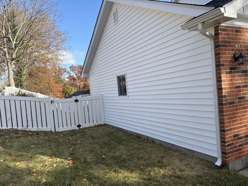 Side wall of a house in white lap siding beside a brick pier, with a white fence and trees