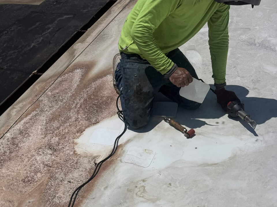 Roofer kneeling on a white single-ply flat roof, holding a square membrane patch and a hot-air welder, with a seam roller and other square patches nearby.
