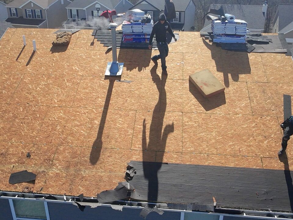 Overhead view of a roof stripped to the deck, with a crew member walking it and shingle bundles staged.