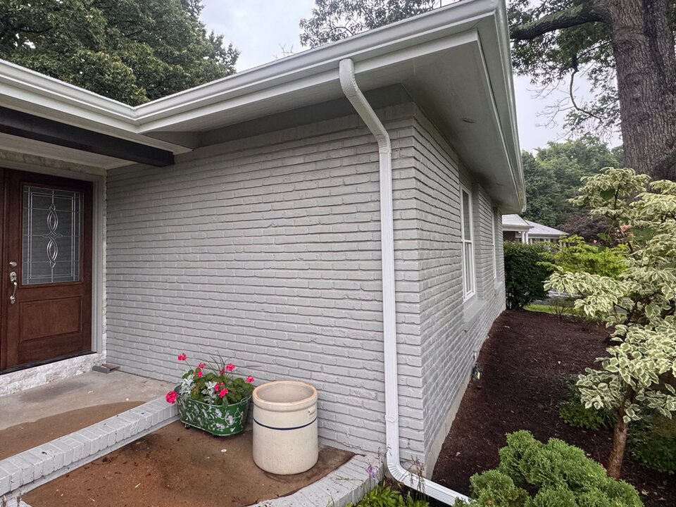 White gutter and downspout on the corner of a light gray brick house, next to an entry door