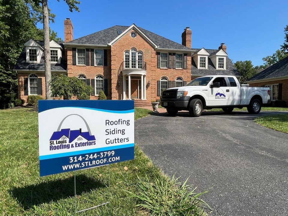 A brick estate home with a St. Louis Roofing & Exteriors yard sign and work truck in the drive.