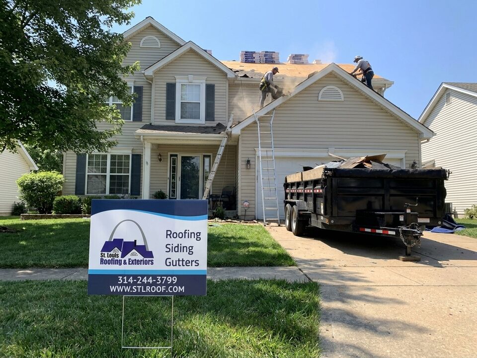 Crew tearing off a roof beside a dump trailer, with a St. Louis Roofing & Exteriors yard sign out front.